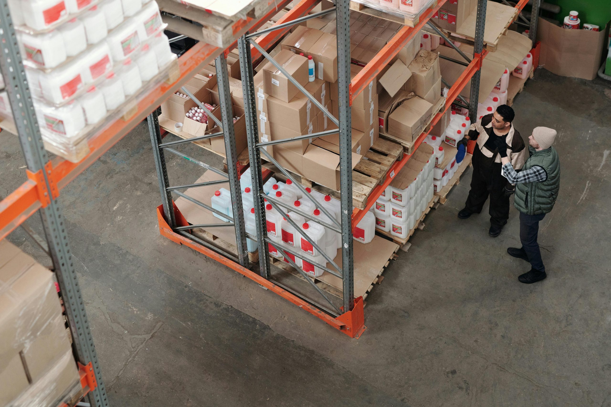 pexels-photo-4481257-4481257 Two warehouse workers discussing logistics beside shelving with boxes and containers.